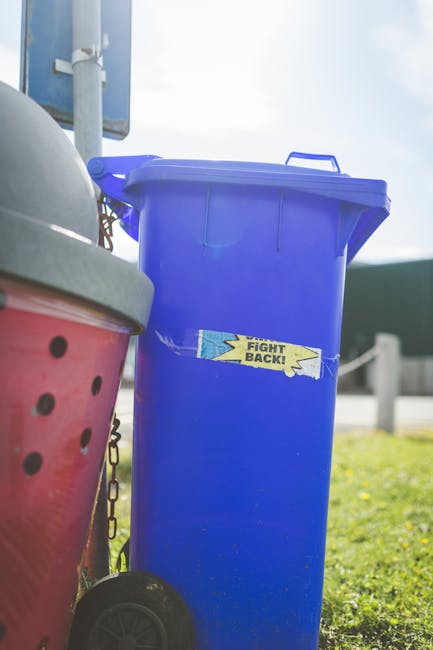 An outdoor scene showing a large cluster of mixed waste and recycling bins overflowing with various types of rubbish, including paper, cardboard, plastic bags, and packaging materials. The rubbish spi